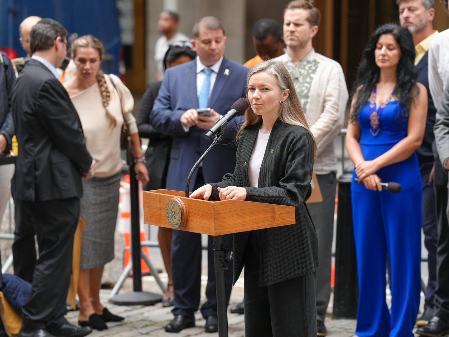 Woman speaking at podium with diverse crowd listening outdoors.